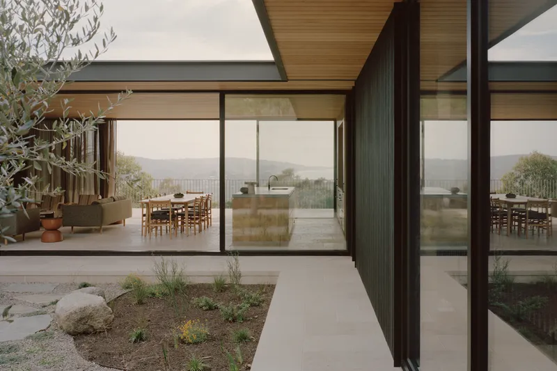 Courtyard pathway between planted beds leading to glazed living space with distant hills visible beyond