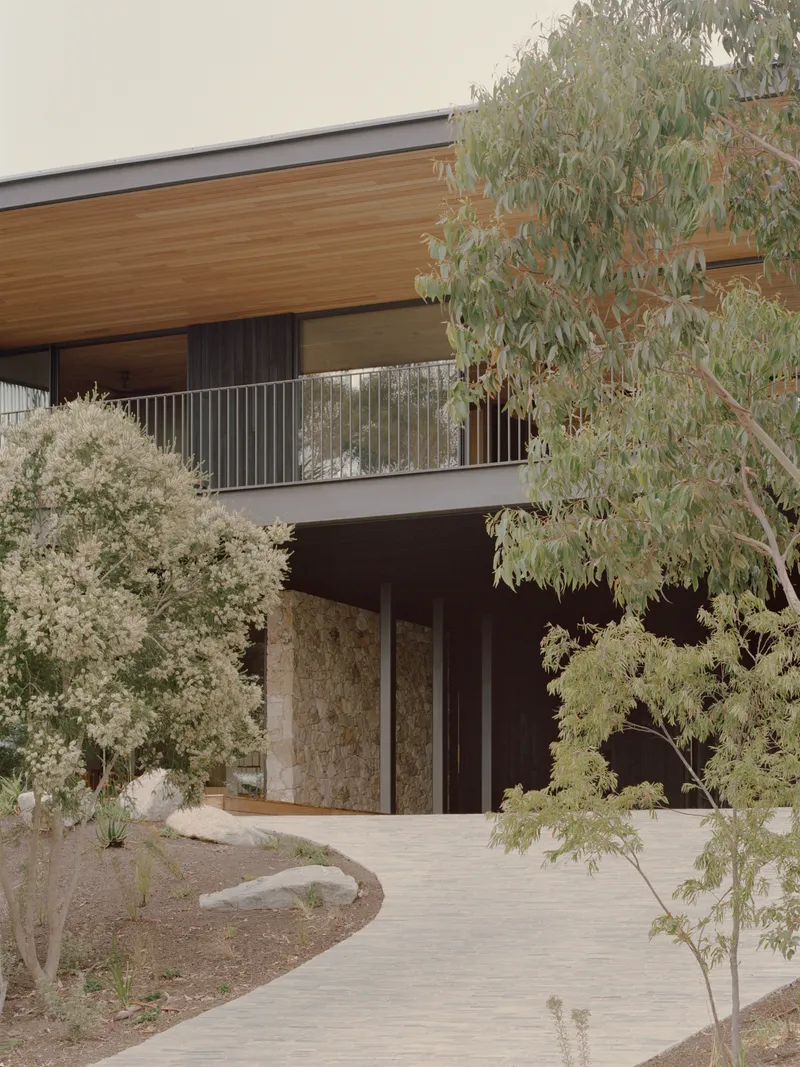 Entry facade with stone-clad walls and timber soffit framed by native plantings and curved pathway