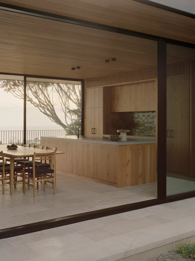 Kitchen island and dining table beneath timber ceiling with ocean view framed by bare tree branches