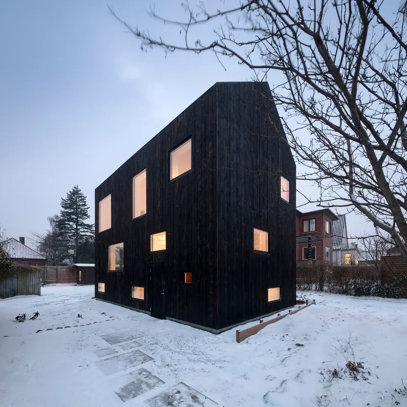 Corner view of the charred timber volume with illuminated windows in a snow-covered yard at twilight