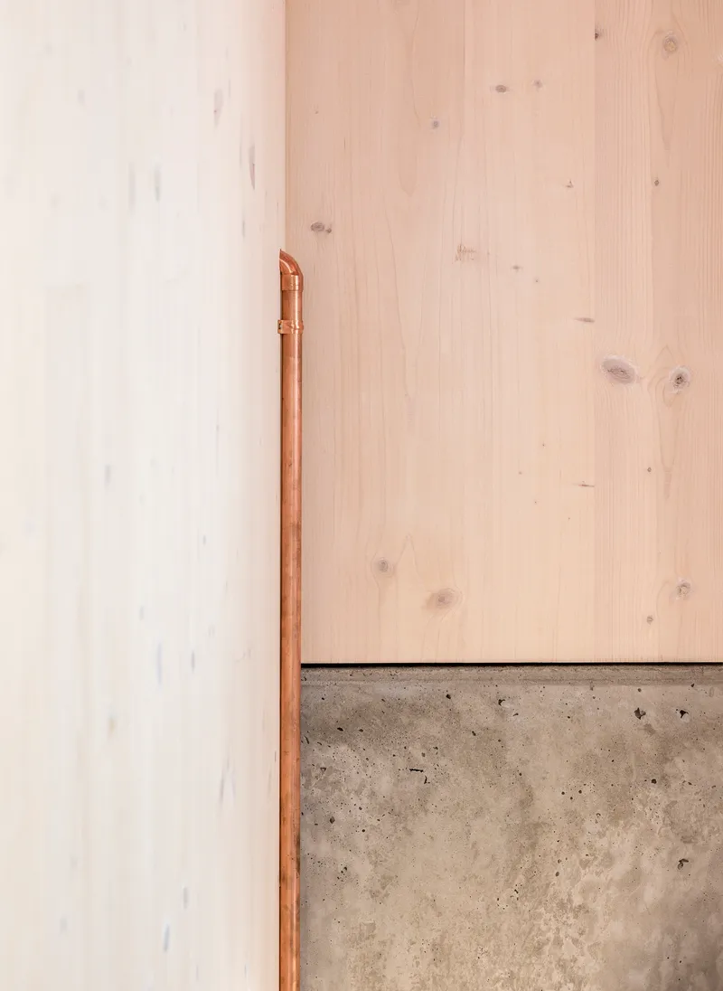 Copper pipe running vertically along white wall above poured concrete plinth and wood panels