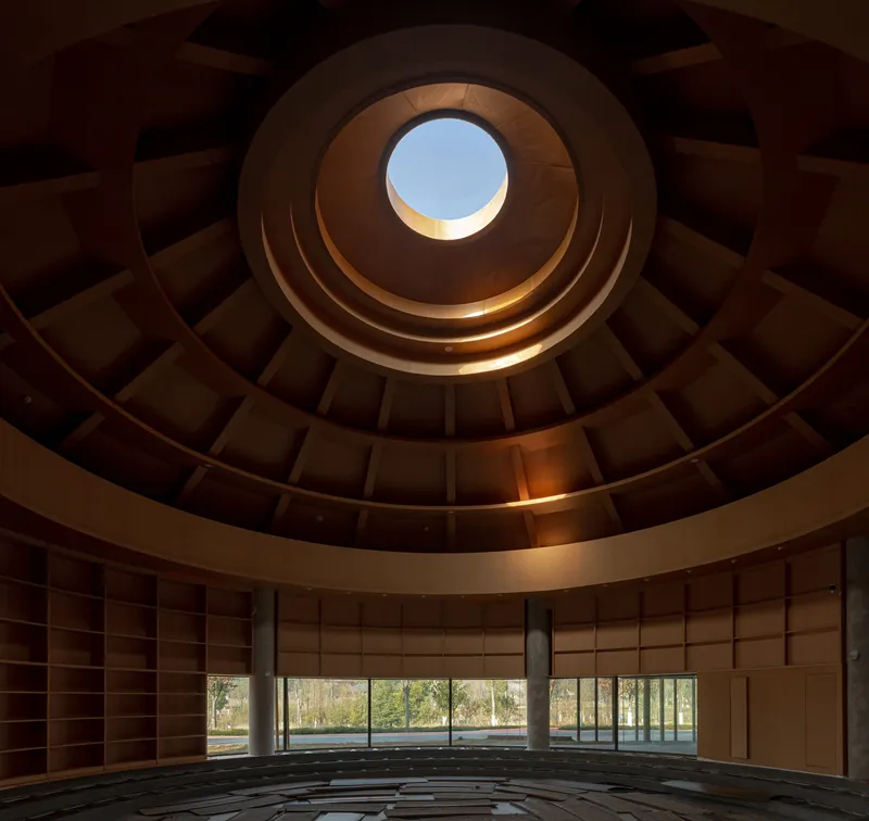 Looking up through a circular coffered dome with concentric timber rings and a central oculus framing blue sky
