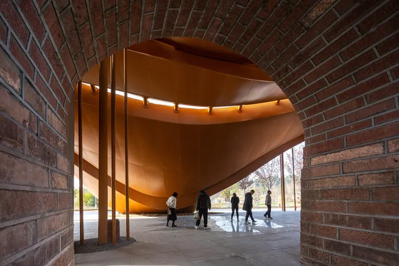 Covered walkway showing a copper-clad volume suspended between red brick walls with visitors beneath