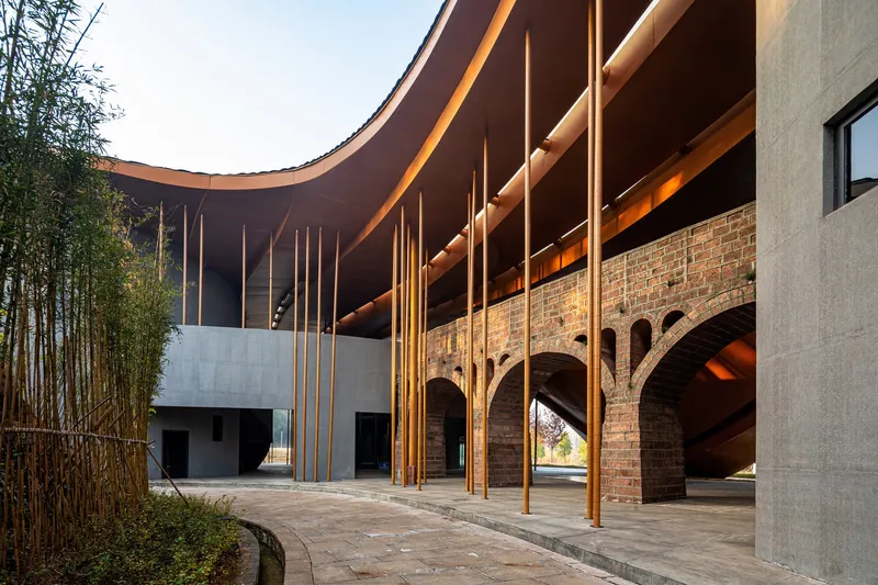Covered courtyard with vertical timber columns beneath the curving roof and arched brick arcade beyond