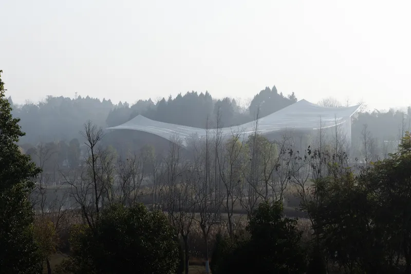 Distant view of the undulating roof form rising above bare trees in heavy mist