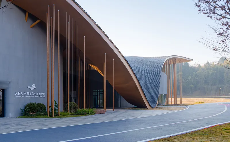 Curved metal roof entry canopy with vertical bronze tubes fronting a paved forecourt