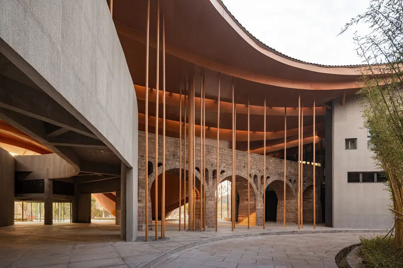 Covered courtyard with exposed brick arches beneath a curving roof and suspended bronze rods at twilight