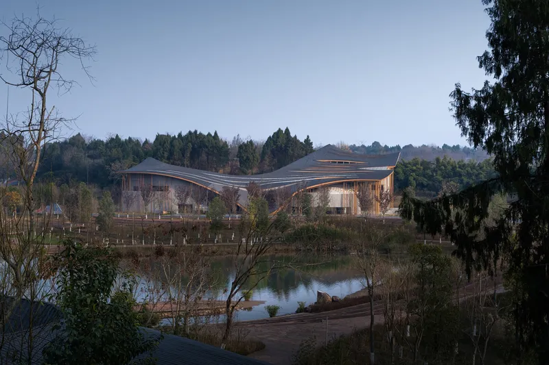Distant view of the wavelike roof set beside a reflective pond and forested hillside