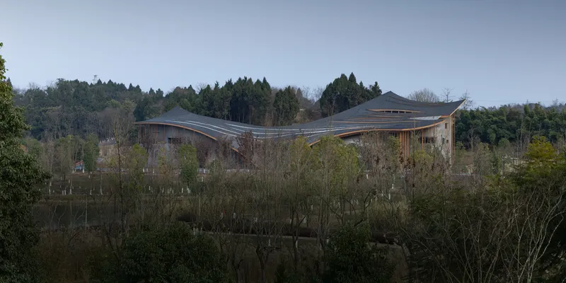 Distant view of the wavelike roof emerging above bare winter trees and pond reflections