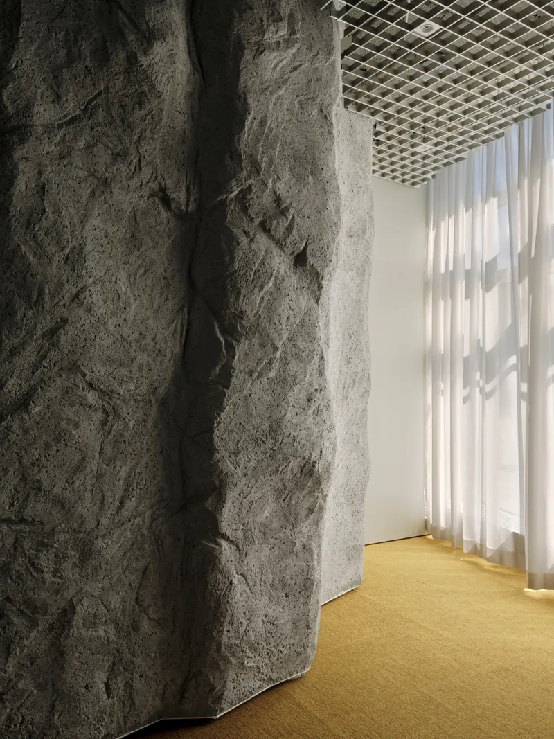 Rough concrete wall edge meeting yellow carpet and sheer curtain under perforated metal ceiling