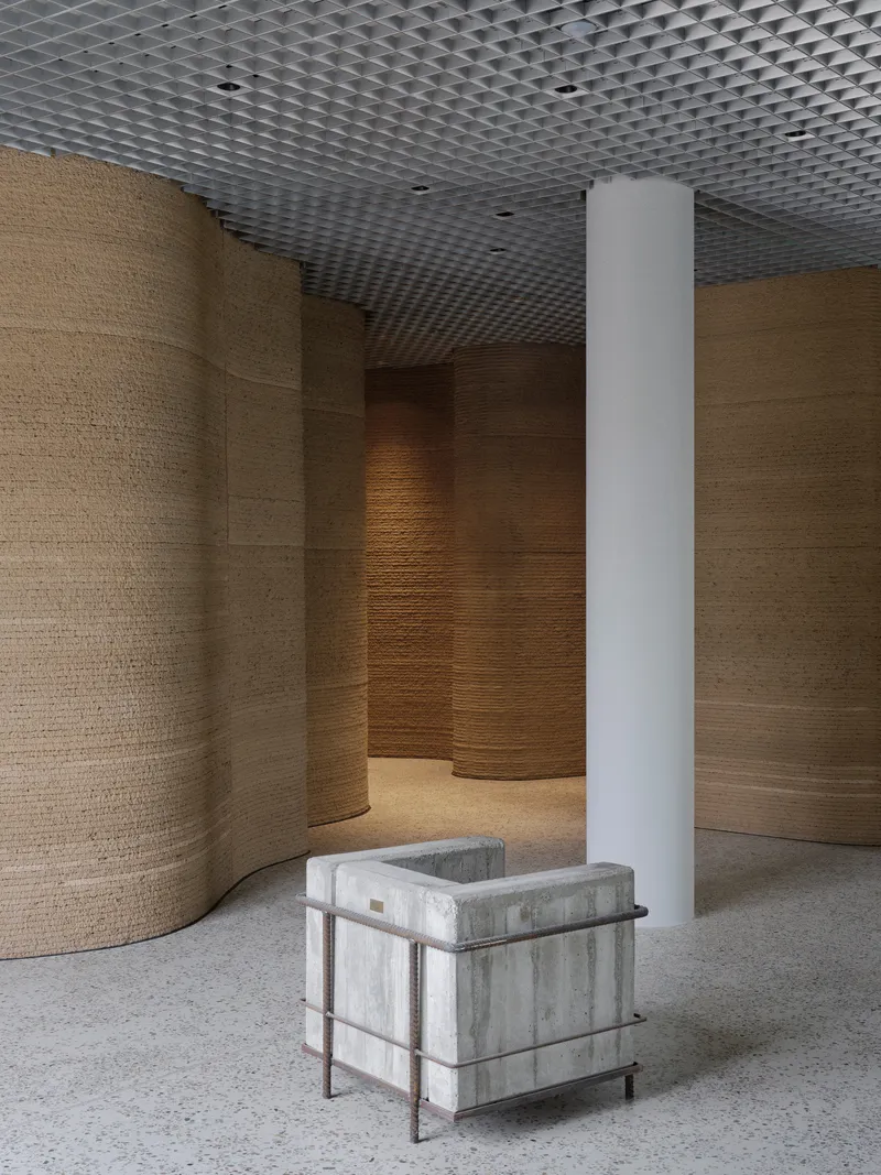 White cylindrical column beside concrete seating cube amid curved timber wall panels and open mesh ceiling