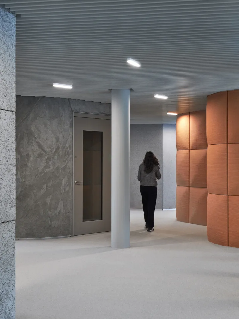 Lobby corridor with ribbed metal ceiling, grey stone walls, copper paneling, and a visitor walking toward a white column