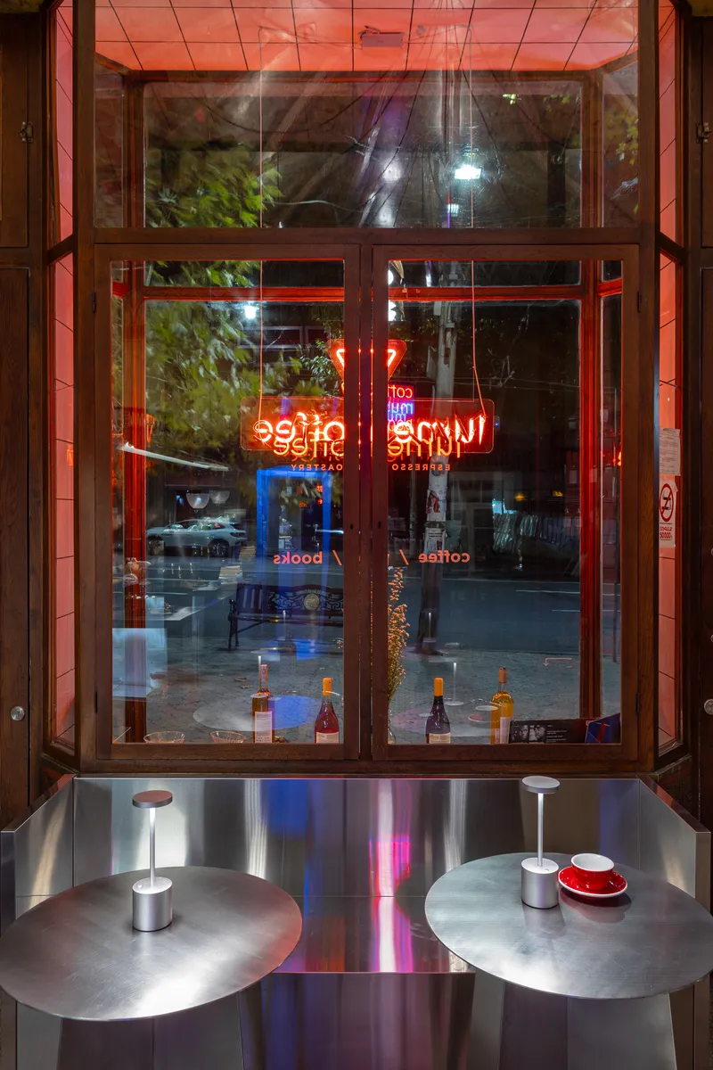 Glass storefront with neon signage reflected in the window above two circular steel tables at night