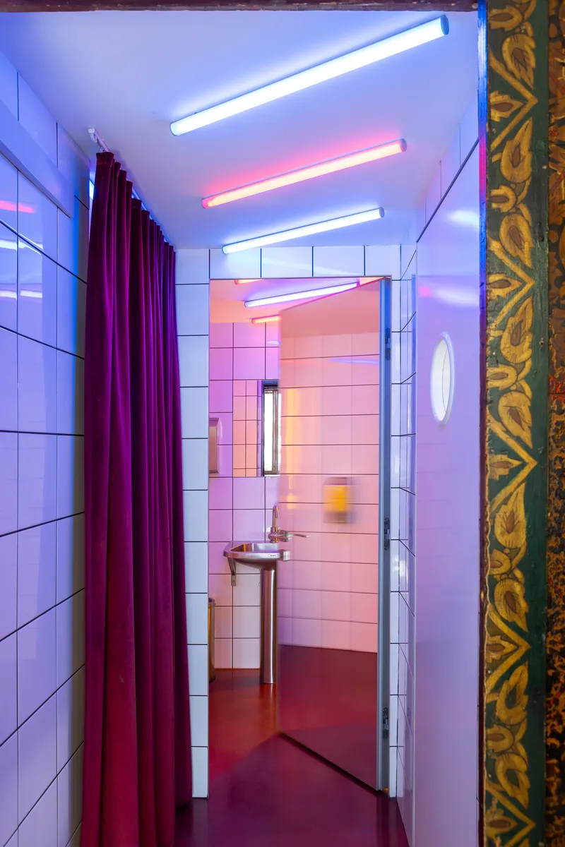 Corridor with white tile walls and coloured neon lighting leading to bathroom with pedestal sink