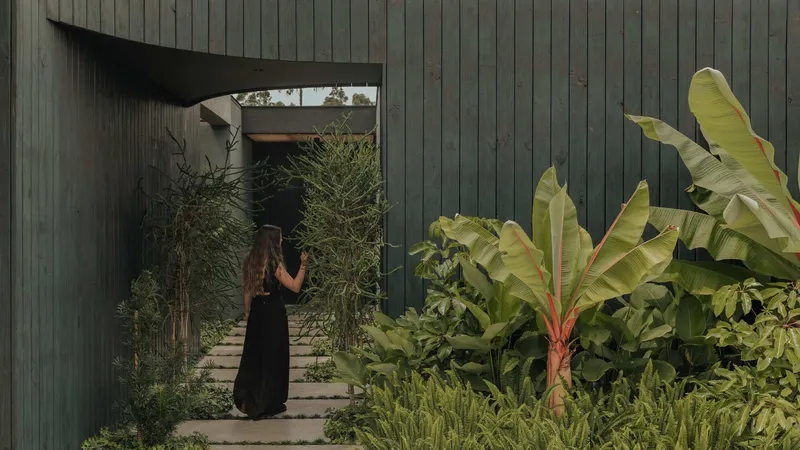 Vertical timber-clad facade with angular opening framing a pathway planted with banana palms and grasses
