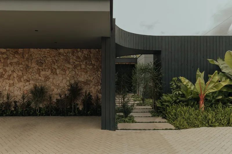 Covered carport with stone wall and curved vertical timber cladding beside planted pathway and banana palms