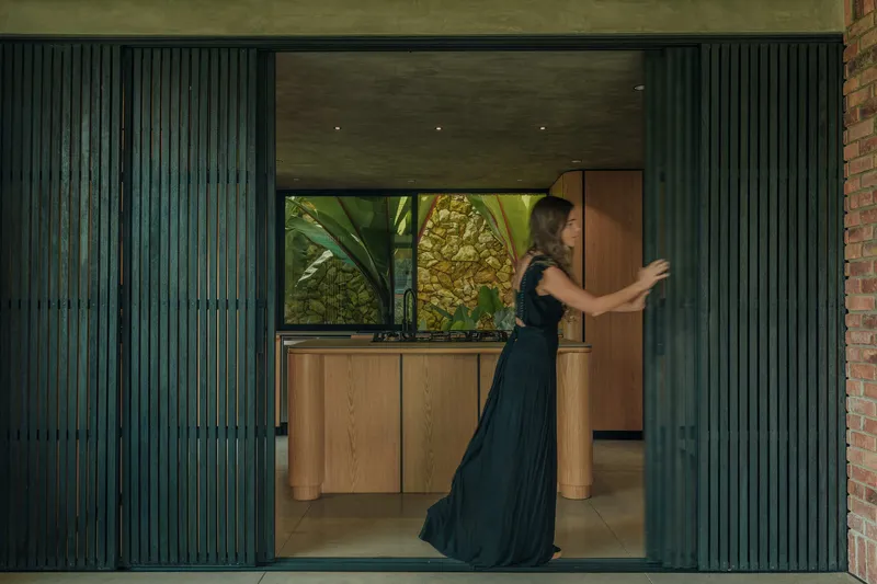 Kitchen view through folding dark green slatted panels as a woman in evening attire opens a timber door