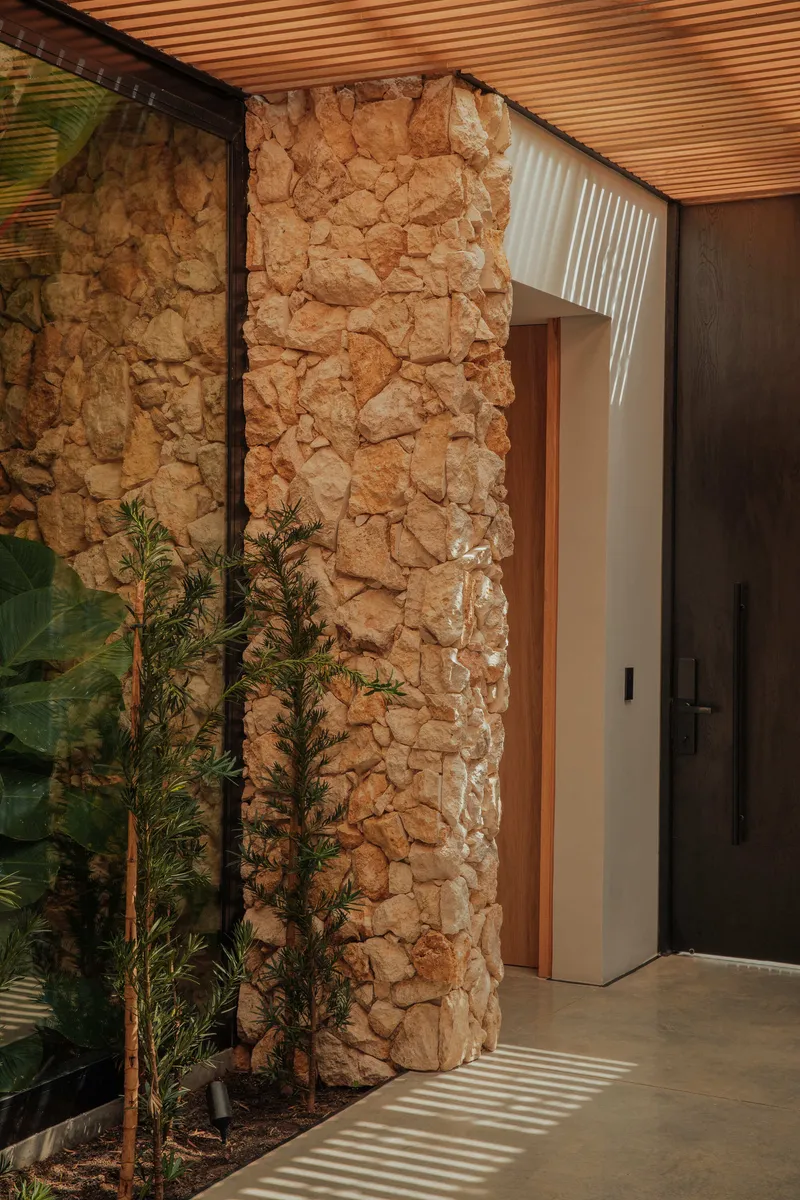 Rough stone wall beside entry door with planted bed and slatted timber ceiling casting linear shadows