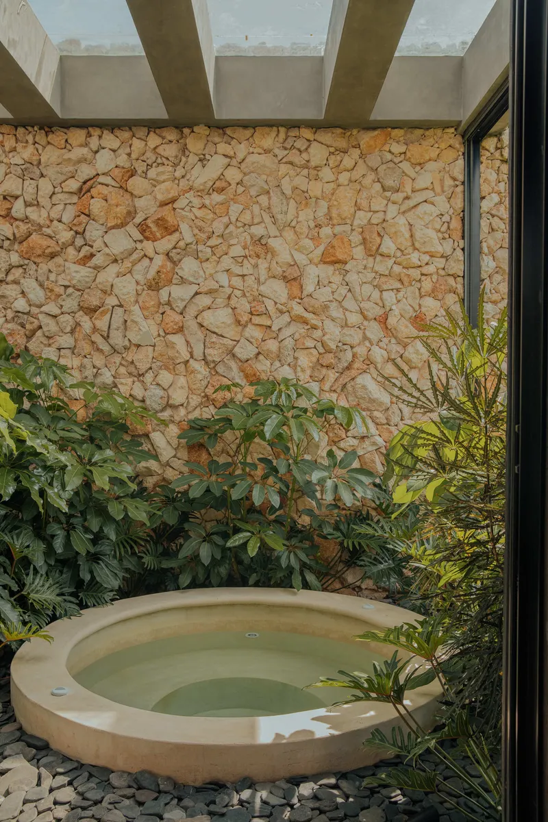 Sunken circular plunge pool in courtyard with rough stone wall and dense planted border under concrete beams