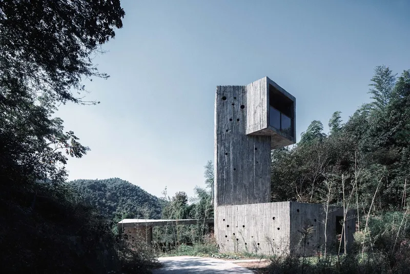 Vertical concrete tower with board-formed walls rising above a wooded hillside road