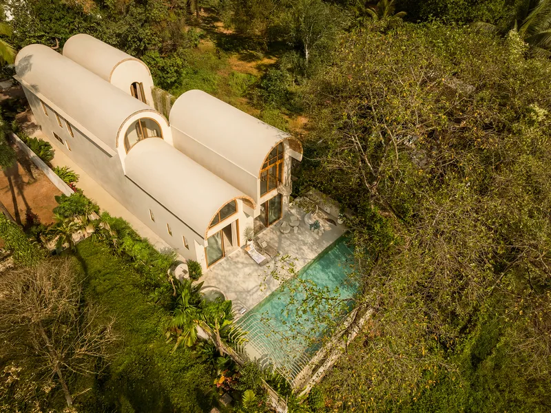 Aerial view of three barrel-vaulted volumes with arched openings beside a pool surrounded by tropical vegetation