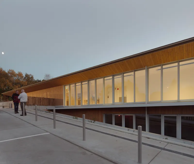 Sloped timber-clad facade with full-height glazing and two figures walking along the exterior ramp at dusk