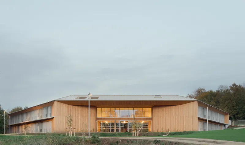 Timber-clad building with folded roof and glazed entry set against lawn and autumn treeline