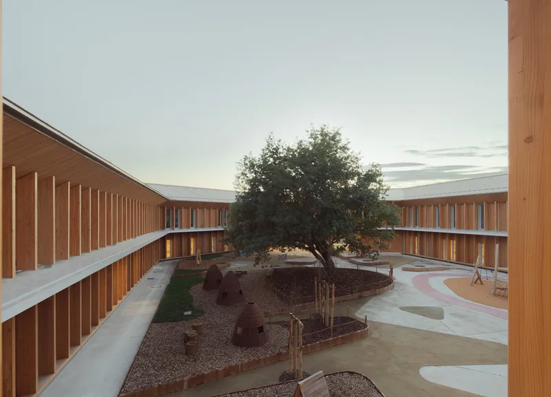Central courtyard at dusk with tree, woven shelters and timber-clad wings under standing-seam metal roofs