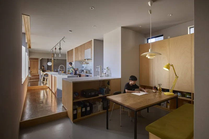 Open kitchen and dining area with light wood cabinetry and a child studying at a desk under pendant lighting