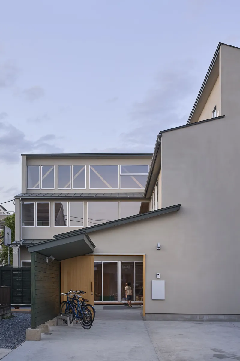 Street entry showing rendered walls and glazed upper volume with bicycles parked under carport