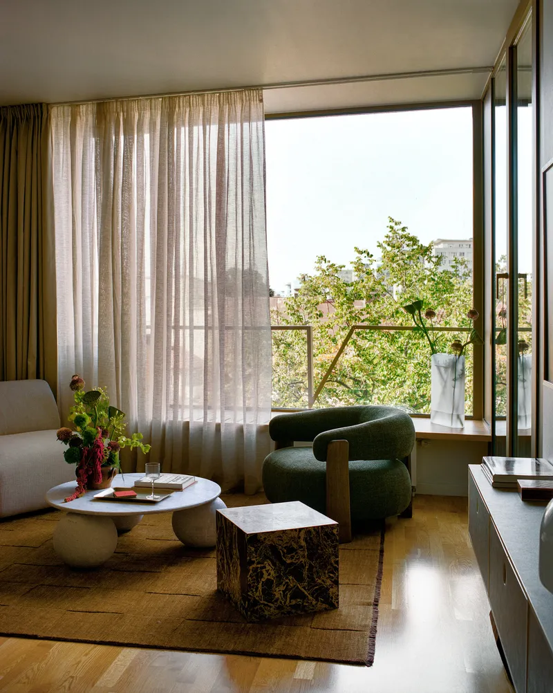 Living room with sheer curtains and open balcony door overlooking treetops and distant buildings