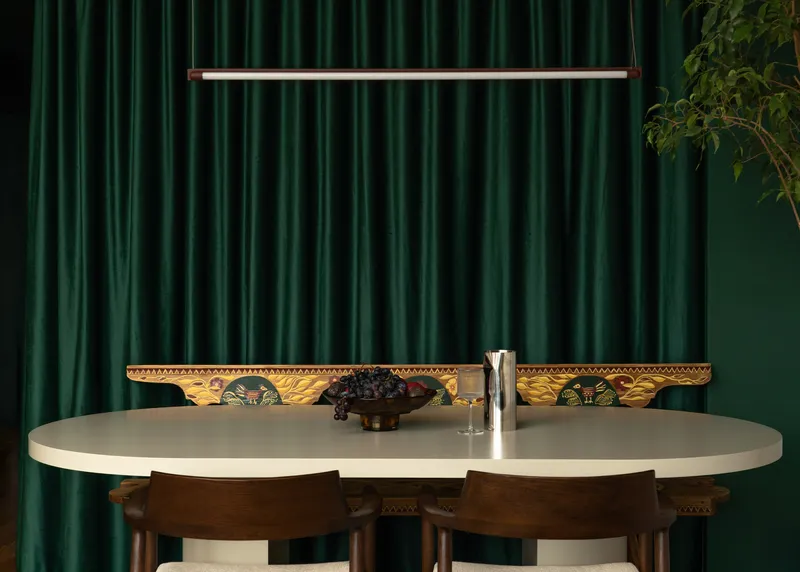 Oval dining table with linear pendant light against deep green velvet curtains