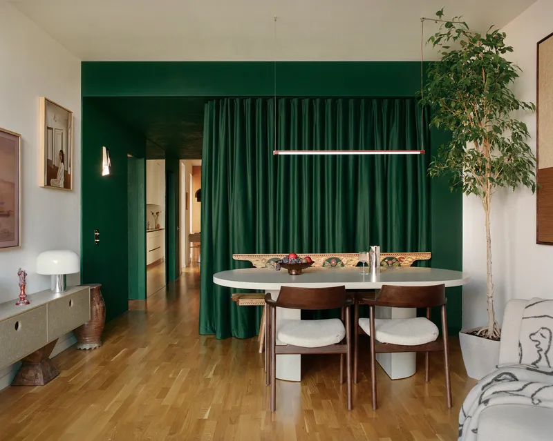 Dining area with green paneled wall and draped curtains beside an indoor tree and timber flooring