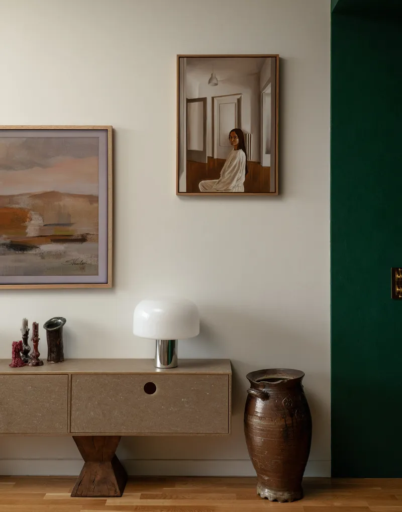 White wall with framed artworks above a timber credenza and ceramic urn near a green door jamb