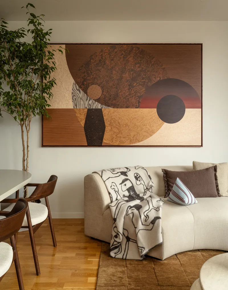 Living area with cream sofa beneath a geometric wall panel and potted plant by timber dining chairs