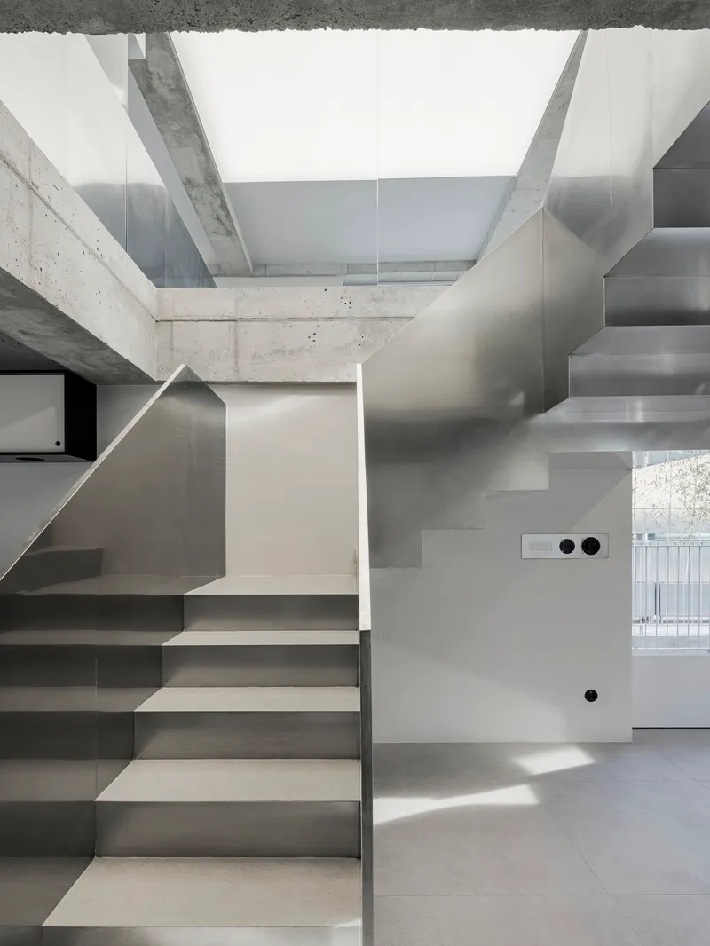 Intersecting stainless steel staircases beneath concrete beams with bright daylight casting sharp shadows