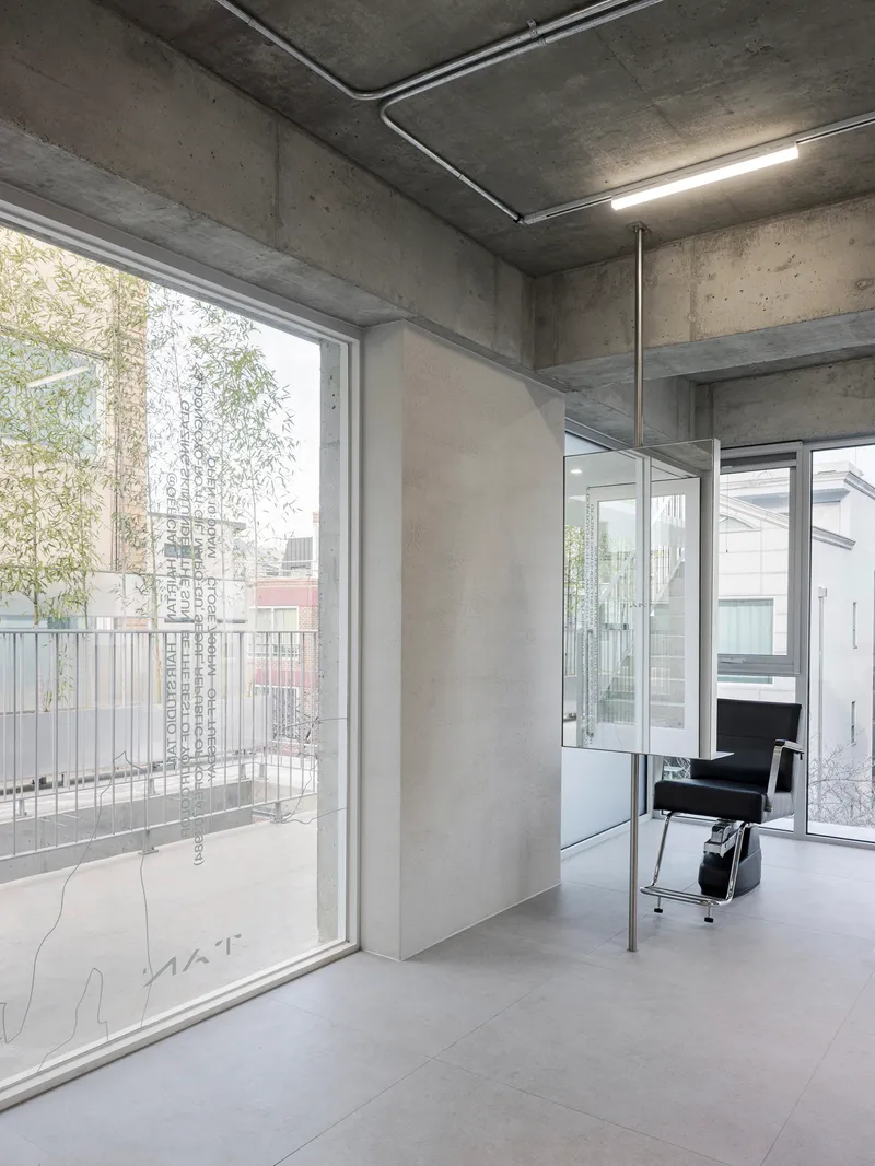 Interior corner with exposed concrete ceiling, large windows opening to bamboo, and a salon chair