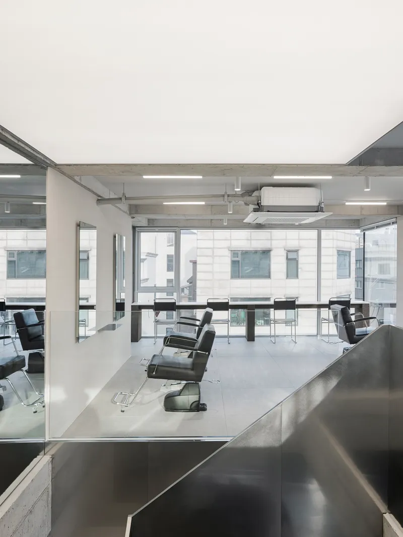Salon floor with rows of barber chairs facing full-height glazing and exposed concrete ceiling