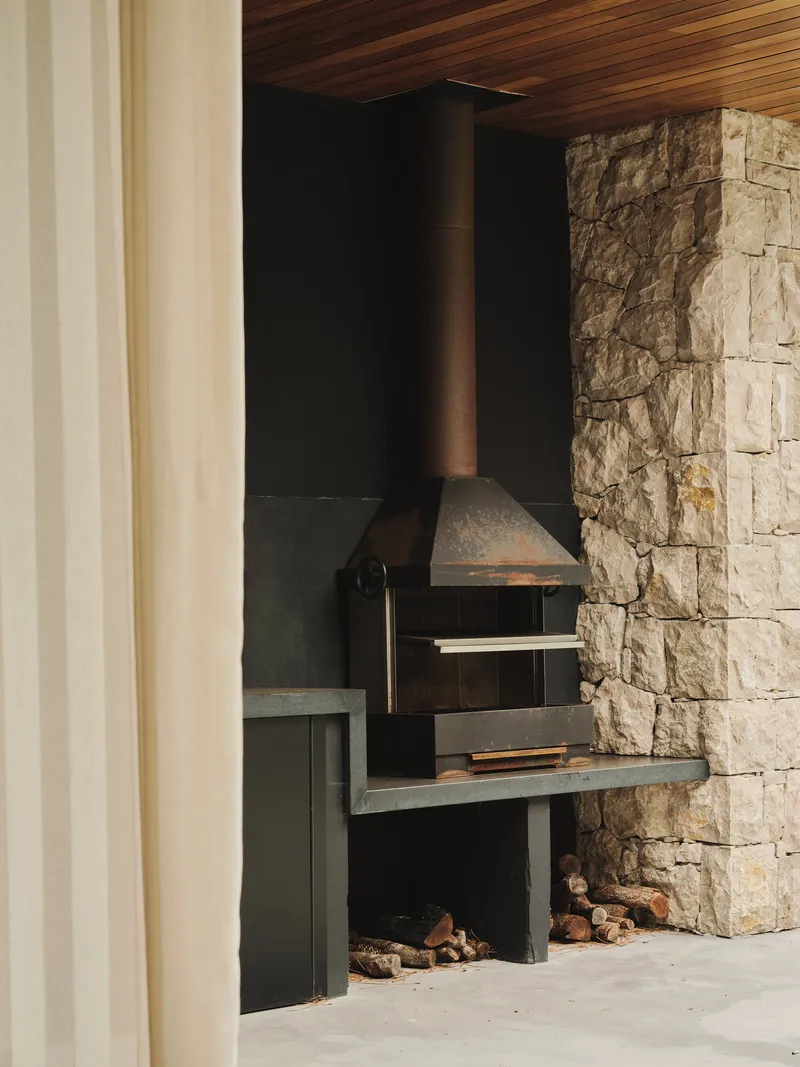 Covered outdoor barbecue area with stone wall, timber ceiling and metal grill under warm afternoon light