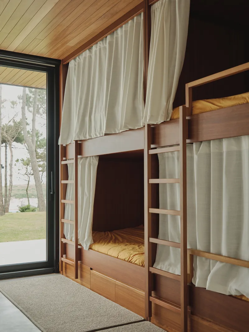 Built-in timber bunk beds with linen curtains and ladder beneath a pitched ceiling with forest views beyond