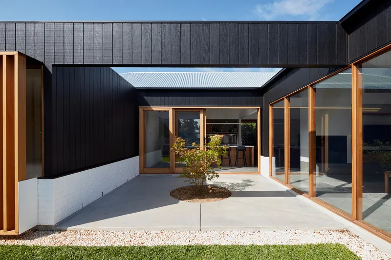 Interior courtyard with concrete paving, a planted tree and timber framed openings under a blue sky