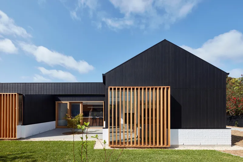 Street view of the black gabled volume with timber screen and white brick base in afternoon sun