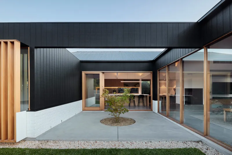 Enclosed courtyard with concrete paving, central planted circle, and black cladding surrounding timber-framed openings