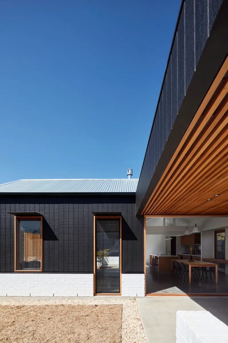 Exterior courtyard view showing charcoal cladding, white brick base, and timber soffit under clear blue sky