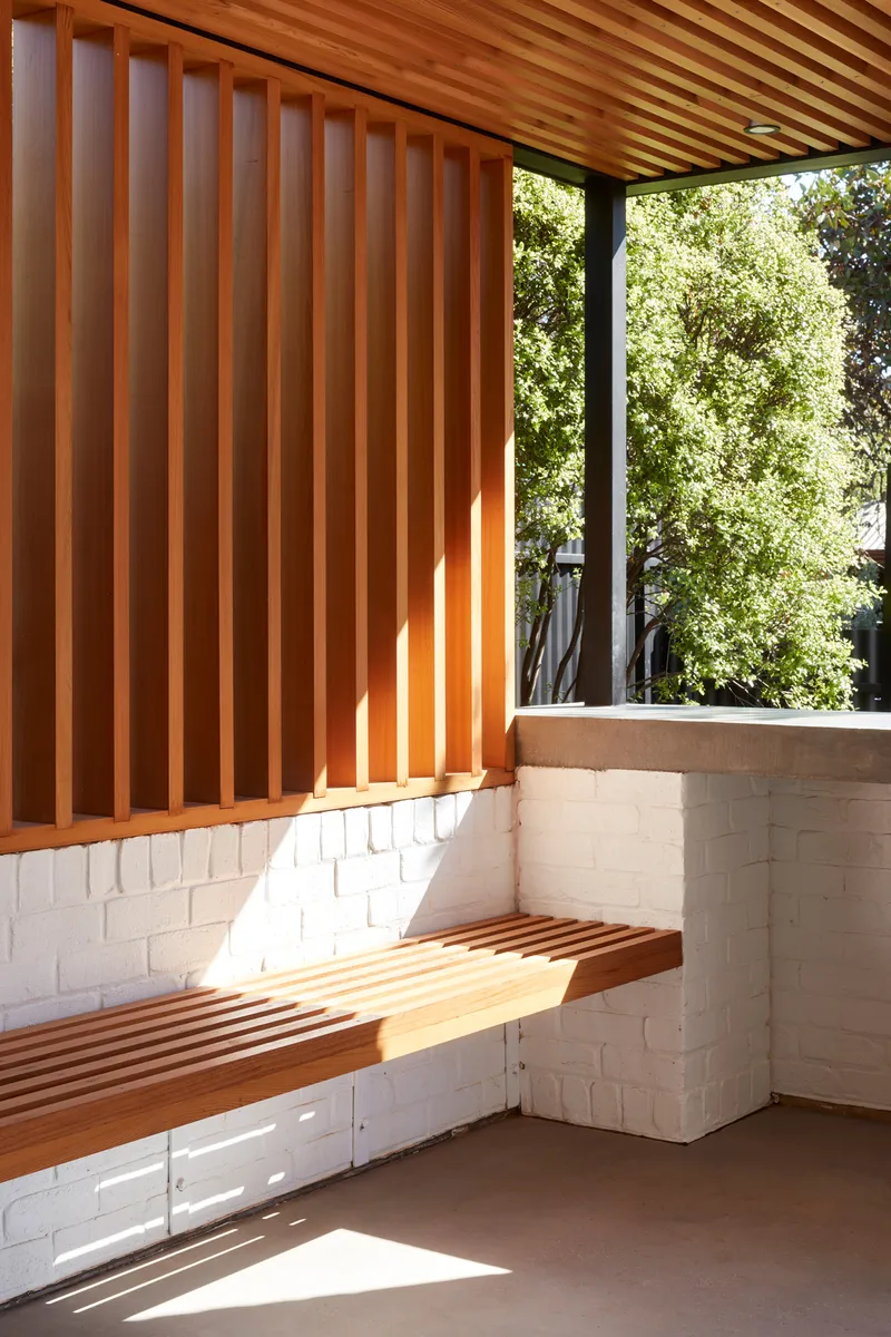 Covered terrace corner with vertical timber battens, white brick base, and slatted bench in dappled sunlight