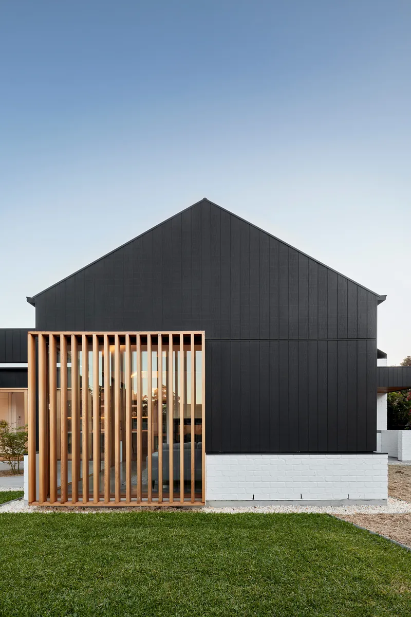 Gabled black timber-clad volume with vertical timber screen over white brick base and lawn