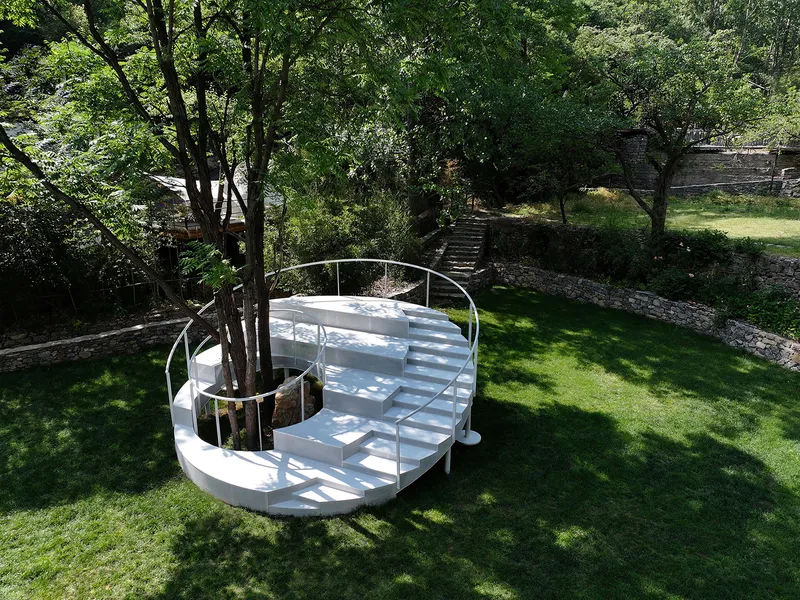 White spiral staircase wrapping around a tree trunk on a lawn in dappled sunlight