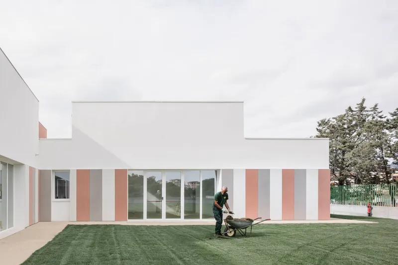Vertically striped facade in white, grey and terracotta panels with a worker pushing a wheelbarrow on the lawn