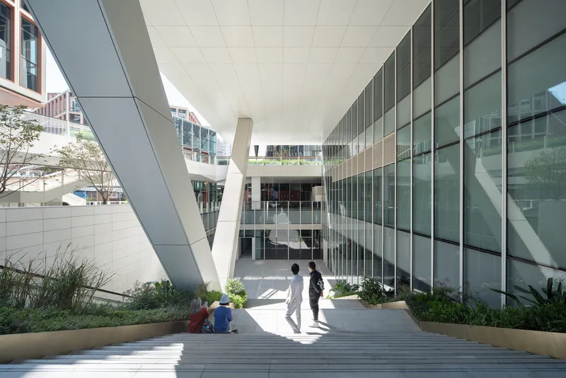 Stepped outdoor seating area beneath angled white columns framing a glazed courtyard with visitors in dappled shade