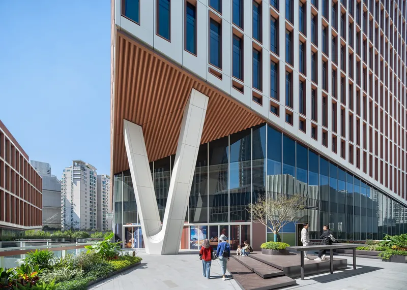 Ground-level plaza with white V-shaped structural columns supporting a ribbed soffit as pedestrians gather near planted beds